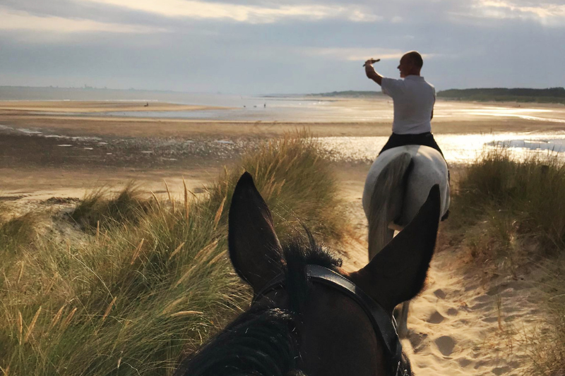 Twee ruiters tijdens een zomerse strandrit. De afbeelding illustreert het belang van een geschikte ondergrond om peesblessures te voorkomen.